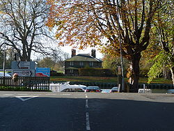  A photograph of the T junction of a road taken from the stalk of the T. A river runs across the top of the T and a small brick cottage can be seen on the far bank.