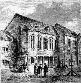 Three two-storey buildings in a staggered row, each with mullioned windows and the central one with a four-columned entrace. Three standing men converse in the courtyard in front, and two men sit at a bench, one drinking and gesturing.