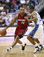 A basketball player, wearing a red jersey with the word «MIAMI» and the number 14 in the front, is dribbling the basketball while another basketball player, wearing a white jersey, attempts to defend him.
