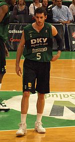 A basketball player, wearing a green-black jersey with the word «DKV JOVENTUT» and the number 5 on the front, stands on a basketball court.