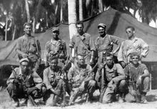 Black-and-white image of ten men in two rows, the top row standing and the bottom row crouching, are all facing the camera. They are wearing military attire and are holding rifles.