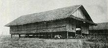 Black-and-white image a grass nipa hut raised a few feet off the ground by wooden supports. Another hut can be seen in the background.