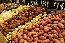 Gros plan sur un étal où les pommes de terre vendues en vrac sont rangées par variétés et couleurs alternées : roses, jaune paille, brun, jaune pailleШаблон:Etc. ; à l’arrière des ardoises en indiquent le prix en dollars par kg.