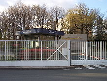 La photographie représente l'entrée de l'entreprise Christian Dior, à Saint Jean de Braye dans le Loiret, en France. Derrière un grand portail blanc qui fait toute la largeur de la photographie, le nom de l'entreprise est inscrit en grandes lettres sur le haut du bâtiment d'accueil, de forme carrée. On distingue également en lettres dorées l'inscription « Parfums Christian Dior » sur un mur isolée face à l'entrée; l'arrière-plan montre l'espace boisé du pôle économique de Charbonnières, dans lequel est implantée l'entreprise que l'on voit au fond derrière les grands arbres.