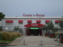 Photographie du Casino de Royan montrant un bâtiment blanc horizontal sur deux niveaux. Le dais marquant l’entrée, les stores de l’étage et l’enseigne sur le toit plat sont de couleur rouge.