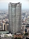 Aerial view of one curved side of a tall building set in front of a cityscape; the facade is symmetrical and divided into three vertical sections: both identical sections on either side are divided by a section in the center that turns into a large atrium window as it nears the building’s roof