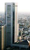 Aerial view of a building’s two white, window-dotted facades set in front of a cityscape; its corners are beveled and are made completely of glass. A brown, horizontal-window-stripped building rising to half of the building’s height is located immediately to its left