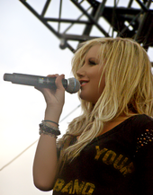 Юная блондинистая певица на сцене поет в микрофон, держа его правой рукой. На ней черная блузка с надписью «Your» и «Band» на ней желтыми буквами и несколько браслетов на правой руке.
