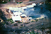 An overhead view from a helicopter of the Mount Carmel Center building. Large columns of smoke are arising from the left side of the building from a fire. One side of the building shows extensive damage. The building is surrounded by dirt paths.