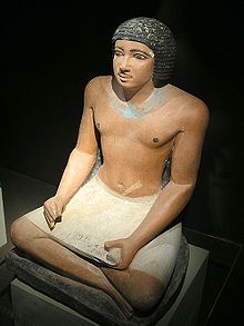 Крашеная, реалистичная, каменная статуя черноволосого мужчины, сидящего со скрещенными ногами и держащего камненное изображение папирусного свитка на коленях