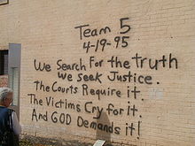 A woman, at the left of the image, is reading a black spray paint message written on a brick wall. The message reads «Team 5 4-19-95 We Search For the truth We Seek Justice. The Courts Require it. The Victims Cry for it. And God Demands it!»