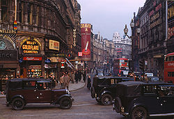 "A busy 1949 city scene in London, England, with black vintage vehicles, red London buses and many pedestrians. Buildings advertise «Wills’s Gold Flake Cigarettes» and «Craven A» cigarettes.