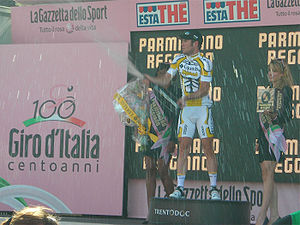 A cyclist popping a champagne bottle on a podium, flanked by two women holding flowers and wine.